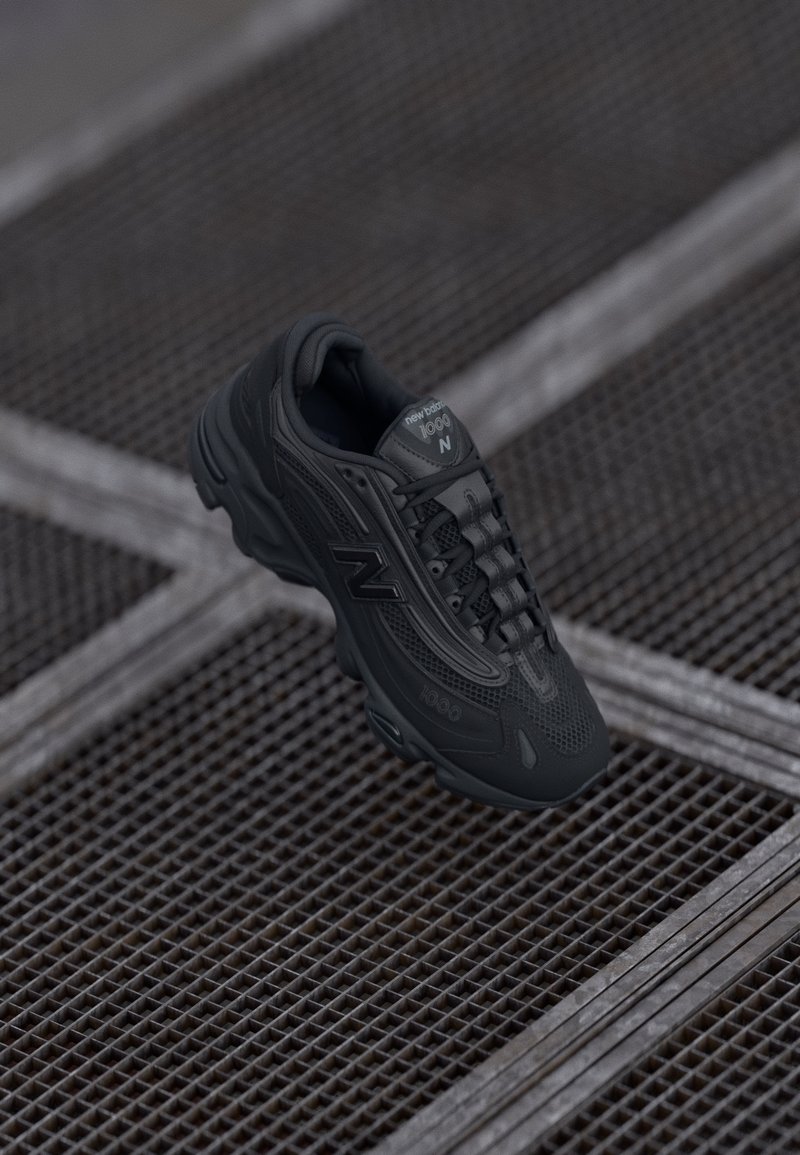 Baskets New Balance 1600 noires suspendues au-dessus d'un sol en grille métallique texturée avec des lignes diagonales formant un motif en grille.