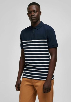 Navyblauwe polo met witte horizontale strepen, 3-knopenknopstrook en geborduurd logo op de borst. Gecombineerd met tan broek.