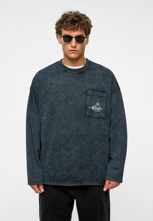 Overmaatse donkergrijze langesleeve shirt van gestructureerde stof, ronde hals en een voorzak met een logo. Draagt het met zwarte broek en een zonnebril.