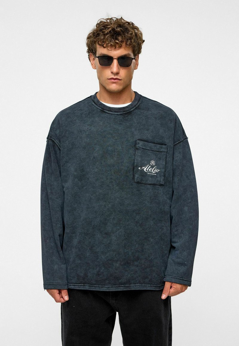 Overmaatse donkergrijze langesleeve shirt van gestructureerde stof, ronde hals en een voorzak met een logo. Draagt het met zwarte broek en een zonnebril.
