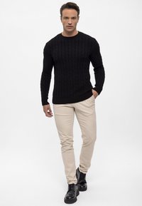 Černý svetr s pleteným vzorem, béžové slim-fit chinos a černé šněrovací boty. Model stojí s jednou rukou v kapse, na čistém bílém pozadí.