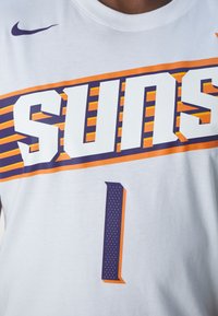 Vit bomulls-T-shirt med "SUNS" i fet lila och orange text, med ett stort "1" nedanför. Nike-logga i lila på vänster axel.