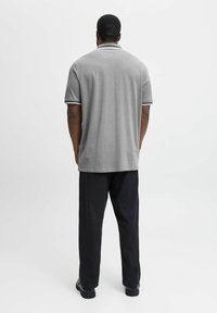 Man staat met zijn rug naar de camera, gekleed in een grijze polo met korte mouwen, zwarte broek en zwarte schoenen, tegen een egale witte achtergrond.