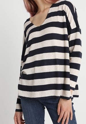 Femme portant un haut ample à manches longues à larges rayures horizontales noires et blanches avec un col en V et un jean bleu.