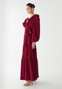 Burgundská maxi šaty s volánovým výstrihom, dlhými puff rukávmi, uterákom na páse a vrstvenou sukňou. Hladká látka a dĺžka po zem.
