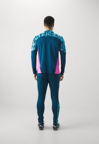 Sportoutfit in Türkis mit pinkfarbener Akzentuierung, geometrischem Muster an den Schultern, taillierter Form und schmal zulaufenden Hosen mit seitlichen Details.