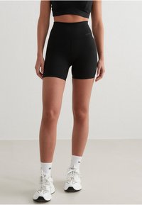 Shorts negros de talle alto hechos de material elástico, con un diseño ajustado y textura suave. Llevados con zapatillas deportivas blancas y calcetines.