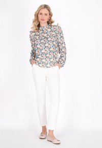 Blomstret bluse med flæser på skuldrene, lyseblå, pink og grønne mønstre. Sammen med hvide slim-fit bukser og lysebeige flade sko med detaljer.