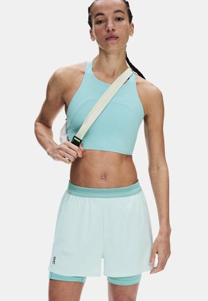 Athletische Frau trägt ein hellblaues Crop-Top und mehrlagige Shorts, hält einen beigen Gurt diagonal über ihrer Brust.