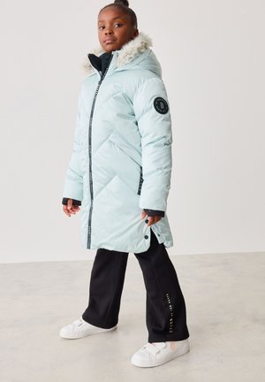 Helles blaues Pufferjacke mit schwarzen Akzenten, einer mit Fell gefütterten Kapuze und Chevron-Steppung. Kombiniert mit schwarzen Hosen und weißen Slippern.