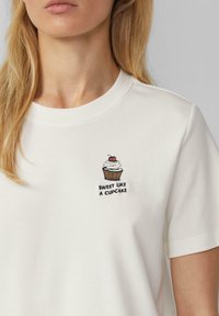 Witte katoenen t-shirt met een cupcake borduursel met een kers bovenop en de tekst "SWEET LIKE A CUPCAKE" eronder in het zwart.