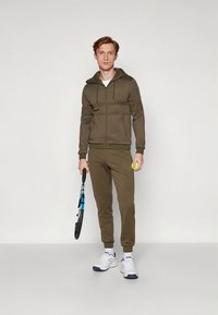 Olíva zöld melegítő szett, sima anyagból készül, cipzáras kapucnis pulóverrel és szűkített szabású nadrággal. Fehér cipővel viselve; teniszkötelet és labdát tart a kezében.