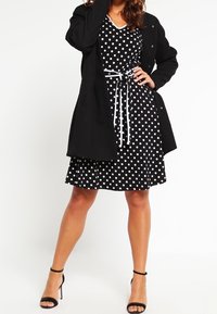 Svart prickig klänning med V-hals och åtsittande midja, matchad med en svart storleksoverdel. Klara klackar fulländar outfiten.