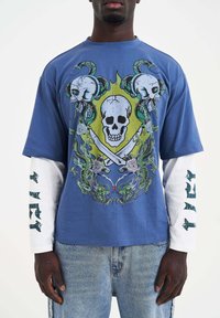 Blaues Grafik-T-Shirt mit Totenköpfen und gekreuzten Schwertern, über einem weißen Langarmshirt mit schwarzem Text. Jeans sind darunter sichtbar.