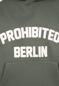 Sudadera verde oliva con letras blancas texturizadas que dicen "PROHIBIDO BERLÍN" encima de un bolsillo frontal.