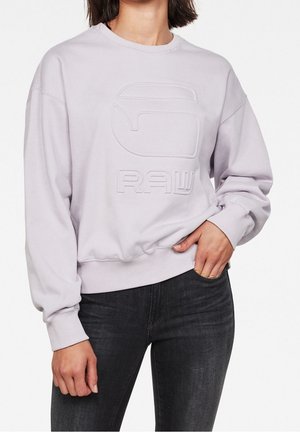 Femme portant un sweat-shirt lavande clair avec le logo "GRAW" en relief et un jean noir ajusté, la main posée sur la hanche.