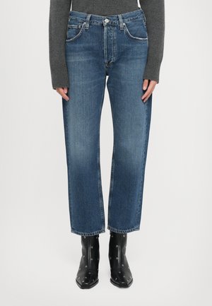 Personne portant un jean court bleu, un pull long gris foncé et des bottines noires avec des œillets en métal, se tenant droit.