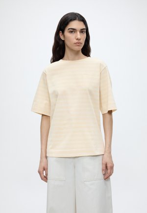 Kvinde med langt mørkt hår iført en løs beige stribet t-shirt og lyse brede bukser, stående mod en enkel hvid baggrund.