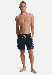 Pantaloni scurți de înot negri cu un model în carouri pe lateral, talie cu șnur și cusături albe. Modelul stă desculț, cu brațele relaxate.