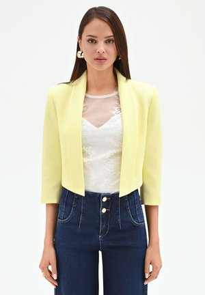 Donna con blazer corto giallo pallido sopra una canotta di pizzo bianca e jeans blu scuro a vita alta, in piedi davanti a uno sfondo bianco.
