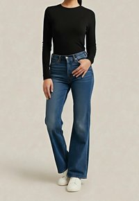 Femme portant un haut noir à manches longues, un jean bleu taille haute et des baskets blanches, debout devant un fond neutre.