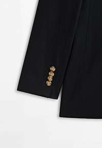 Manche de veste noire sur mesure avec quatre boutons décoratifs marron, présentant une texture de tissu lisse et des lignes nettes et précises.