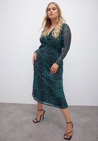 Durchsichtiges türkisfarbenes Kleid mit schwarzem Leopardenmuster, langen Ärmeln und einem geknoteten Design vorne. Kombiniert mit schwarzen Riemchen-Heels.