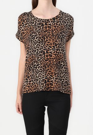 Frau in einem kurzärmeligen Top mit Leopardenmuster und rundem Ausschnitt, kombiniert mit schwarzen Hosen, steht vor einem weißen Hintergrund.