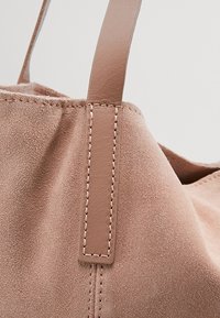 Sac en daim blush avec une bandoulière en cuir lisse. Couture soignée le long du bord de la bandoulière. Texture douce avec un design souple.