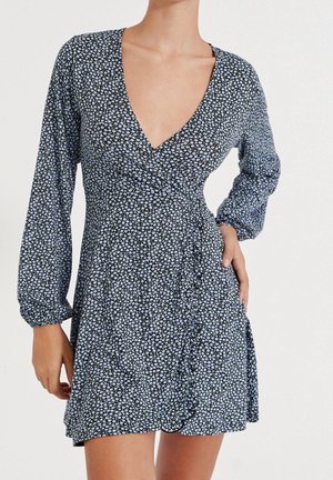 Woman wearing a black wrap dress with long sleeves and a small blue floral pattern, posing with one hand on hip and one arm relaxed.