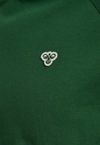 Zaļš kokvilnas krekls ar balto izšūto bišu logotipu kreisajā pusē, ar gludu tekstūru un klasisku apaļu kakla dizainu.