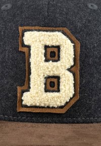 Filzkappe mit einem großen, cremigen, strukturierten Buchstaben "B" in braunem Rand auf einem dunkelgrauen Hintergrund und einem Schirm aus braunem Wildleder.