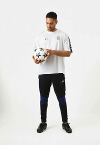 Balta trumpo rankovės marškiniai su logotipu, juodos kelnės su mėlynomis detalėmis, laikantis juodai baltą futbolo kamuolį, dėvint juodus futbolo batus.