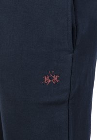 Granatowe spodnie dresowe z miękkiego materiału, ozdobione małym czerwonym haftowanym logo przedstawiającym dwóch jeźdźców na koniach. Posiadają boczne kieszenie.