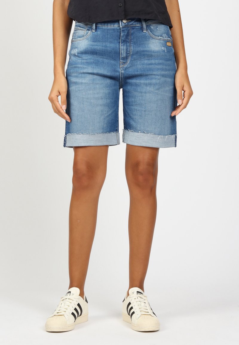 Denim bermuda shorts in light blue wash, featuring a rolled hem, front pockets, and a distressed finish. Paired with white sneakers.