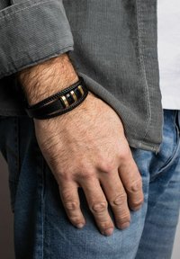 Fekete bőr karkötő, fonott mintával, három arany és fekete fém kiegészítővel, egy kézen viselve, amely egy textúrált szürke pólóra támaszkodik.