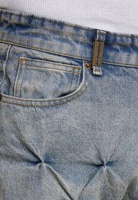 Jasnoniebieskie dżinsy o zniszczonym wyglądzie, z kontrastowymi pomarańczowymi przeszyciami i metalowym guzikiem. Zawierają małą metkę na pasie.