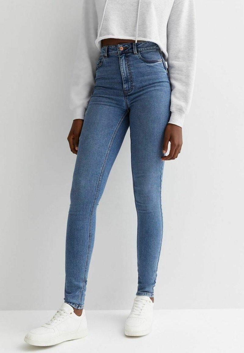 Vartaloon istuvat, korkeavyötäröiset siniset farkut joustavasta denimmateriaalista, sileä pinta, istuva malli ja kontrastisaumaukset, yhdistetty valkoisiin tennareihin.