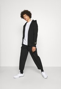 Schwarze Zip-Up-Hoodie mit Kapuze, kombiniert mit einem weißen T-Shirt und schwarzen Hosen. Weiße Turnschuhe und ein Ring runden den Look ab.