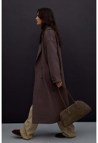 Długi brązowy płaszcz o podwójnym zapięciu, w połączeniu z jasnobeżowymi spodniami i ciemnozieloną torebką. Gładki materiał i wyrazista sylwetka.