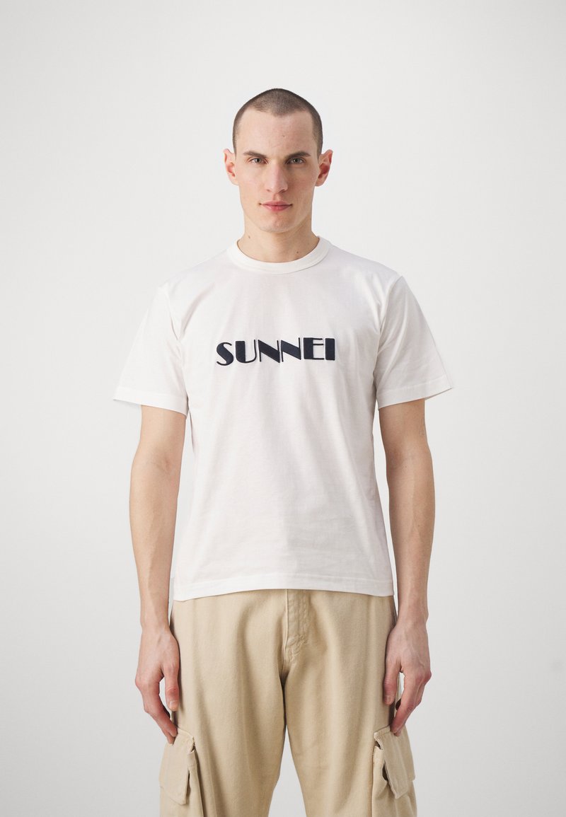 T-shirt alb din bumbac, cu mâneci scurte, decorat cu un logo negru "SUNNEI" pe piept. Poartă pantaloni cargo bej.