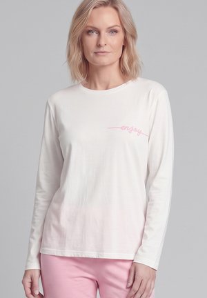 Blonde Frau trägt ein weißes Langarmshirt mit dem pinken Text „"enjoy"“ und hellrosa Hose vor einem einfarbigen grauen Hintergrund.