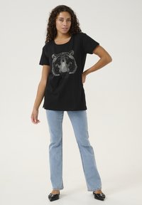 T-shirt en coton noir avec un graphique détaillé d'ours argenté. Coupe à col rond et manches courtes, associé à un jean bleu clair.