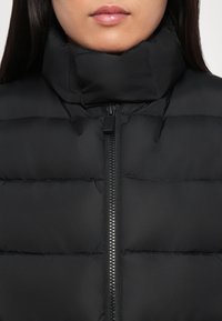 Femme portant une doudoune noire avec un col montant et une fermeture éclair visible à l'avant, lèvres fermées et cheveux raides et foncés visibles sur les côtés.