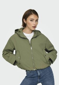 Veste à capuche verte avec fermeture éclair, dotée de poches latérales et d'une coupe décontractée. Portée par-dessus un t-shirt blanc et associée à un jean bleu.