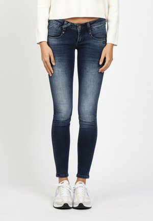 Woman wearing dark blue skinny jeans, white cropped top, and white Nike sneakers, standing against a plain light background.