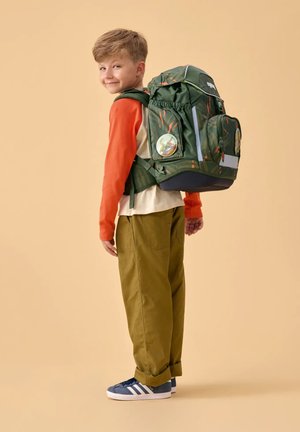 Young boy wearing an orange and beige shirt, green pants, and blue sneakers carrying a large green backpack with dinosaur patches.