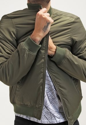 Olivgrüne Bomberjacke mit durchgehendem Reißverschluss, geripptem Strickkragen und -ärmeln sowie glattem, glänzendem Stoff. Darüber liegt ein gemustertes Hemd.