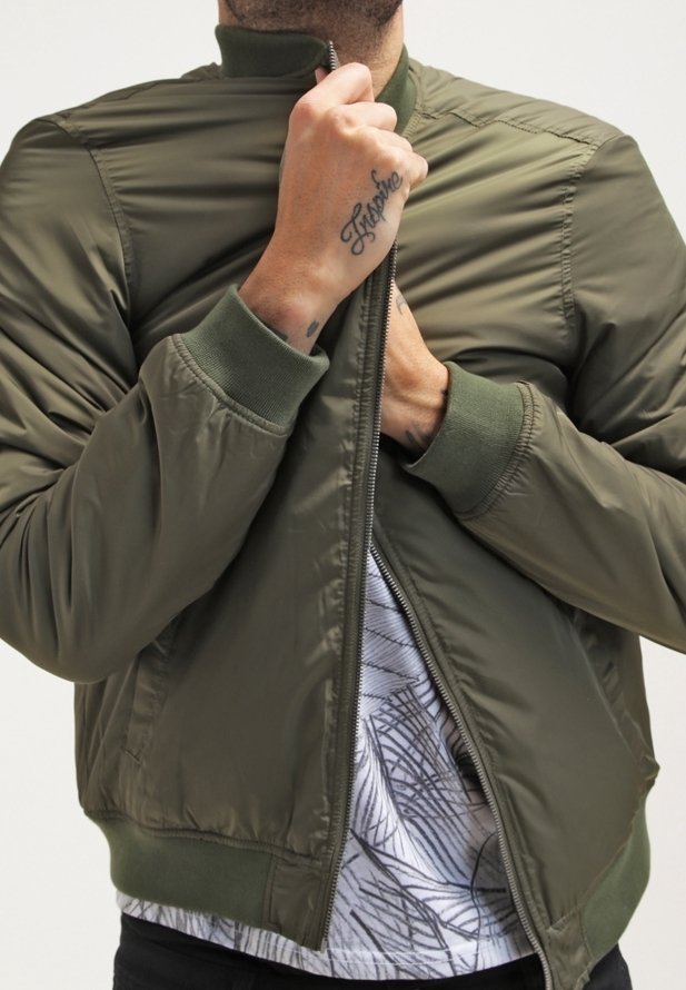 Kurtka bombka w kolorze oliwkowej zieleni z zamkiem z przodu, prążkowanym dzianinowym kołnierzem i mankietami oraz gładką, błyszczącą tkaniną. Nałożona na wzorzystą koszulę.