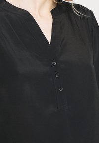  Närbild av en person som bär en svart V-ringad blus med tre synliga svarta knappar längs en central knäppning.